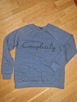 Blå sweatshirt från Amisu med text - Säljer en blå sweatshirt från Amisu i storlek XS. Tröjan har ett upphöjt blommönster i tyget och svart texten 'Completely in love' framtill. Mjuk mudd vid ärmslut och nederkant, perfekt för chill dagar.
