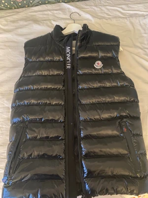 Svart dunväst från Moncler - Säljer en svart, glansig dunväst från Moncler med dragkedja framtill. Västen har två fickor med dragkedja och Moncler-logga på bröstet. Klassisk quiltad design och stilren look, perfekt för lager-på-lager under kyliga dagar.