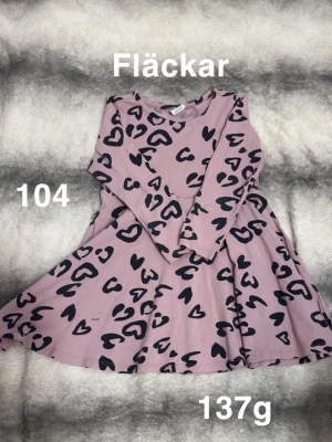 Rosa klänning med hjärtmönster - Supersöt rosa klänning med svarta hjärtan över hela tyget. Klänningen har lång ärm och en snurrig kjol som ger extra rörelse. perfekt för lek och vardag. Passar dig som gillar gulliga detaljer och lekfull stil.🌸 Fläckar. Storlek 104