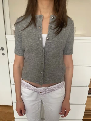 Grå kortärmad kofta från Zara - Kortärmad grå kofta från Zara i mjukt stickat material. Koftan har rund hals och har snygga silverfärgade knappar framtill. Perfekt att slänga över ett linne för en chill och stilren look. Passar både till vardags och till finare tillfällen.                  Bara använd fåtal gånger och har inga defekter eller fläckar. 