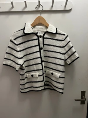 Randig kofta från H&M Divided - Kortärmad kofta från H&M Divided i vitt med svarta horisontella ränder. Tröjan har en klassisk krage, knappar framtill och två dekorativa fickor med knappdetaljer. Ribbstickade kanter vid ärmslut, krage och nederkant.❣️❣️