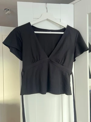Svart omlottopp från Gina Tricot - Svart omlottopp från Gina Tricot i mjuk bomull. Toppen har v-ringning, korta ärmar och knytband i midjan som ger en snygg siluett. Perfekt till jeans eller kjol för en stilren look. Inte så stretchig 🩷