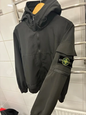 Svart vindjacka från Stone Island - Säljer en svart vindjacka från Stone Island med ikonisk patch på ärmen. Jackan har huva, dragkedja framtill och praktiska fickor. Materialet är polyester och modellen har en clean, sportig look som passar perfekt till streetwear.