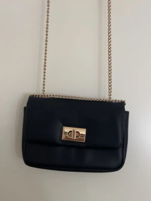 Svart axelväska från H&M med kedja - Stilren svart axelväska från H&M med guldfärgad kedjerem och vridlås framtill. Väskan är i slätt material och har ett innerfack med dragkedja. Perfekt storlek för det viktigaste och enkel att matcha till olika outfits.