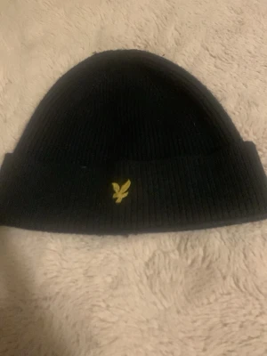 Svart ribbad mössa från Lyle & Scott - Säljer en svart ribbstickad mössa från Lyle & Scott med deras klassiska gula fågelbrodyr framtill. Mössan har uppvikt kant och är riktigt mjuk och skön. Perfekt accessoar för kyliga dagar.