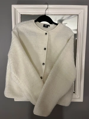 Vit teddyjacka från 157, storlek S - Mysig vit teddyjacka från 157 i storlek S. Jackan har rund halsringning, tryckknappar framtill och två fickor. Perfekt för dig som gillar en enkel men trendig look. Materialet är fluffigt och ger en skön vibe till alla outfits.