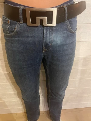 Svart skärp i skinn från J.Lindeberg - Säljer ett svart bälte från J.Lindeberg i äkta skinn, tillverkat i Italien. Skärpet har ett brett band och en stor, fyrkantig metallspänne med modern design. Perfekt accessoar för att lyfta din outfit.