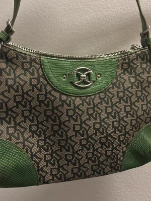 Grön och beige axelväska från DKNY - Snygg axelväska från DKNY med gröna detaljer, och beige tyg med svart logomönster. Väskan har silverfärgad metallogo framtill och dragkedja upptill. 