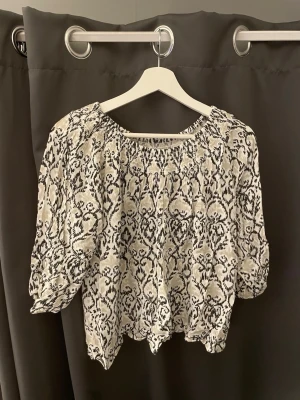 Svartvit blus med mönster - Säljer en luftig svartvit blus med unikt mönster och rundad hals. Blusen har trekvartsärm med lätt puff och resår vid ärmslutet. Materialet är tunt och känns skönt mot huden, perfekt för dig som gillar en avslappnad men ändå snygg stil.