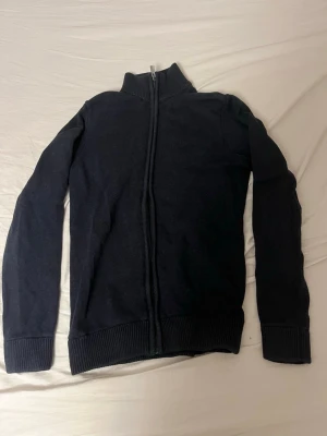 Mörkblå stickad kofta från Dobber - Säljer en mörkblå stickad kofta från Dobber med hel dragkedja framtill och hög ribbad krage. Tröjan har långa ärmar och ribbade muddar vid ärmslut och nederkant. Perfekt för lager på lager under kyliga dagar.