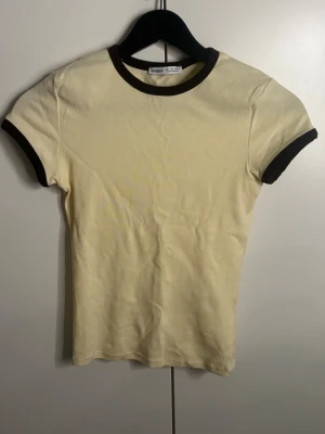 Gul t-shirt med kontrastkant från Bershka - Säljer en ljusgul t-shirt från Bershka med mörk bruna kontrastkanter vid halsringning och ärmslut. Modellen är figurnära och har korta ärmar. Perfekt till jeans. Tröjan har använts ungefär två gånger💗