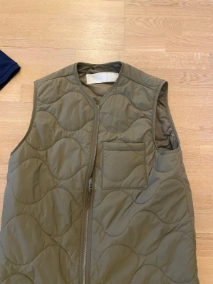Olivgrön quiltad dunväst från ARKET - Säljer en olivgrön quiltad dunväst från ARKET med vågigt mönster, dragkedja framtill och en bröstficka. Västen har en clean look och är perfekt att slänga över en hoodie eller tröja för extra värme.