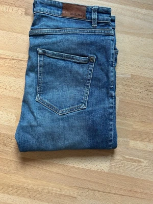 Blå jeans från Dobber, storlek 30 - Säljer ett par klassiska blå jeans från Dobber med snygga detaljer som metallknappar och logga på fickan. Jeansen har fem fickor och normal passform. Perfekta till vardagslooken och funkar året runt.