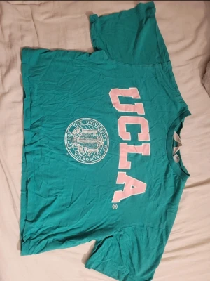 Turkos UCLA t-shirt med tryck - Säljer en turkos t-shirt från UCLA med stort vitt och rosa tryck framtill. Oversized passform och klassisk rund hals. Perfekt för dig som gillar college-vibe och vill ha en chill look.