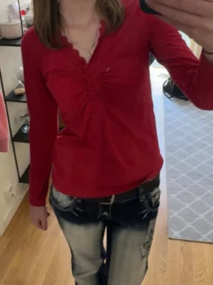 Röd blus med spets från KappAhl - Snygg röd blus från KappAhl i storlek S med lång ärm och v-ringad hals. Blusen har dekorativ spets runt halsringningen och knappar framtill. Materialet är mjukt och lätt, perfekt för dig som gillar detaljer och färg.