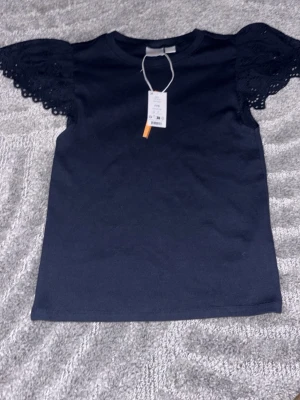 Marinblå topp med spetsärmar Gina Tricot - Supersnygg marinblå topp från Gina Tricot med korta, vida spetsärmar som har broderade detaljer. Klassisk rund halsringning och rak passform. Perfekt att styla med jeans eller kjol för en trendig look.