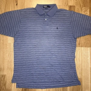 Gråsvart randig pikétröja Polo Ralph Lauren - Klassisk blå pikétröja från Polo Ralph Lauren i storlek XL enligt sizetaggen. Den sitter mer som en stor M Tröjan har tunna vita och mörkblå ränder, krage och två knappar framtill. Ikoniska broderade loggan på bröstet. Perfekt till jeans eller shorts för en chill look.
