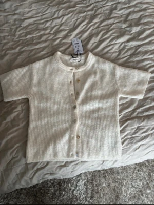 Vit kortärmad kofta från Gina Tricot - Kortärmad kofta från Gina Tricot i offwhite. Ribbstickad med rund hals och knappar framtill. Tillverkad i en mjuk ullblandning som känns både trendig och mysig. 