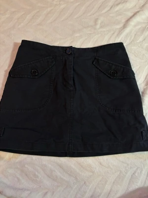 Svart kortkjol från H&M, strl 36 - Säljer en svart kortkjol från H&M i storlek 36. Kjolen har coola fickdetaljer med knappar framtill och en ficka bak. 