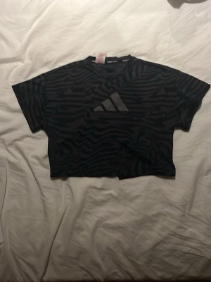 Svart mönstrad Adidas crop top - Säljer en svart crop top från Adidas med zebramönster och stor logga framtill. T-shirten har korta ärmar och en boxig passform, perfekt till träning eller streetwear. Materialet känns mjukt och lätt mot huden.