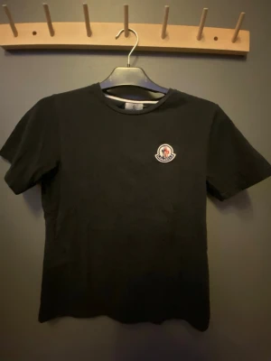 Svart Moncler t-shirt med logga - Svart t-shirt från Moncler i klassisk modell med rund hals och korta ärmar. På bröstet sitter Monclers ikoniska broderade märke. Hör av dig om det intresserar dig!