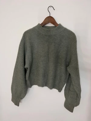 Grå stickad croppad tröja - Mysig grå-grön stickad tröja med croppad passform. Tröjan har en högre ribbad halsringning. Säljer då jag inte använder så mkt längre ❣️