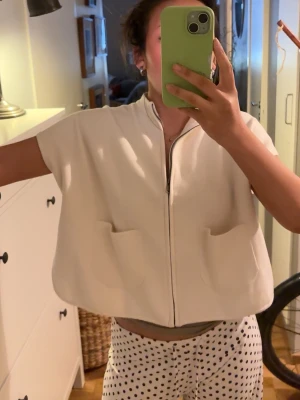 Beige kortärmad kofta från Zara - Kortärmad beige kofta från Zara med dragkedja framtill och två stora fickor. Avslappnad passform och mjukt material, perfekt för lager-på-lager. Enkel och stilren design som funkar till många outfits. Barnstolrke men sitter som en s på mkg