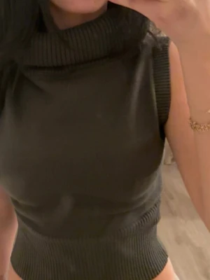 Mörk grön stickad polotopp - Säljer en mörkgrön stickad polotopp med bred ribbad krage och ärmlös design. Toppen har en croppad passform och är perfekt för att styla med höga jeans eller kjol. Materialet är mjukt och stretchigt vilket gör den superbekväm.