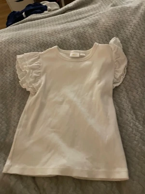 Vit topp med volangärm från Gina Tricot - Supersöt vit topp från Gina Tricot i ribbad bomull med korta volangärmar i broderad spets. Super skön tröja på sommaren eller under något eller vanligt. Använd någon enstaka gång på sommaren men annars inte super fin med axlarna rekommenderar verkligen på sommaren. Pris kan andras på bra förslag.