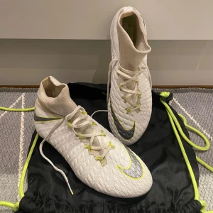 Nike Hypervenom Elite SG - Snygga och sparsamt använda vita fotbollsskor från Nike med snörning och neonfärgade detaljer. Skorna har en strömlinjeformad design med en slitstark sula och är perfekta för spel på gräsplaner. Inga hål eller separationer.