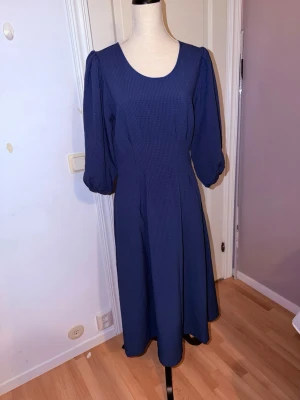 Marinblå långklänning med trekvartsärm - Snygg marinblå långklänning med rund halsringning och trekvartsärmar. Klänningen har en lätt strukturerad yta och markerad midja som ger en smickrande siluett. Perfekt för dig som gillar enkel men stilren design.