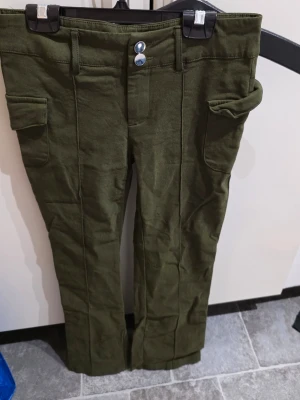 Olivgröna cargobyxor med fickor - Snygga olivgröna cargobyxor med raka ben och stora sidofickor. Byxorna har dubbla knappar i midjan och breda hällor för bälte. Tillverkade i ett mjukt och bekvämt tyg som passar perfekt till en avslappnad stil.