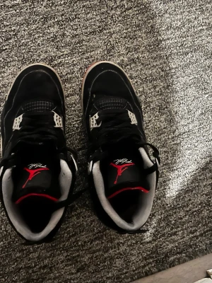 Air Jordan 4 Retro Black/Red - Säljer ett par klassiska Air Jordan 4 Retro sneakers i svart mocka med röda och grå detaljer. Skorna har meshpaneler, plastdetaljer och synlig Air-sula. Ikonisk Jumpman-logga på plösen och bekväm vadderad insida. Perfekta för dig som gillar streetwear och sneakerskultur. Fick dem i present, använd några gånger.