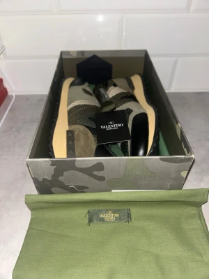 Valentino Garavani - Snygga sneakers från Valentino med camomönster i olika nyanser av grönt, beige och svart. Skorna har detaljer i mocka och läder, samt klassiska nitar på sidan av sulan. Kommer i originalkartong och med dustbag. Perfekta för dig som vill sticka ut med stil. Skokartongen har lite skada vid hörnen.