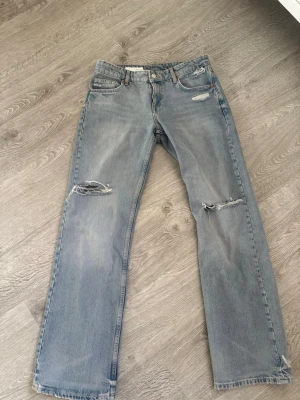 Ljusa raka jeans med slitningar Zara - Säljer ett par ljusblå raka jeans från Zara med slitna detaljer och hål på båda knäna. Jeansen har klassisk femficksmodell och är tillverkade i denim. Perfekta för en avslappnad och trendig look.