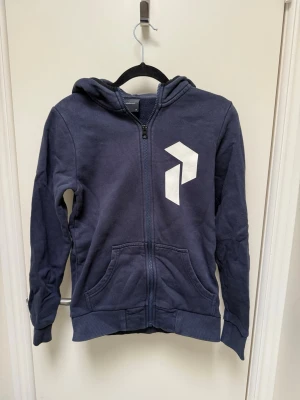 Peak performance Mörkblå hoodie med vit logga - Mörkblå peak performance hoodie med dragkedja och stor vit grafisk logga på bröstet. Tröjan har huva, två fickor framtill och ribbade muddar vid ärmar och nederkant. Tillverkad i mjukt bomullsmaterial, perfekt för en avslappnad stil.