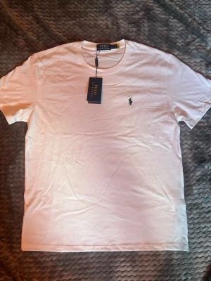 Vit t-shirt från Polo Ralph Lauren - Klassisk vit t-shirt från Polo Ralph Lauren med den ikoniska broderade loggan på bröstet. T-shirten har rund hals och korta ärmar, tillverkad i mjuk bomull för en skön och stilren look. Perfekt basplagg för alla tillfällen.