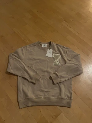 Beige sweatshirt från Ami Paris - Snygg beige sweatshirt från Ami Paris med deras ikoniska hjärta-A-logga i vitt på bröstet. Tröjan har rund hals, ribbade muddar och är tillverkad i mjuk bomull. Perfekt för en clean och avslappnad stil.