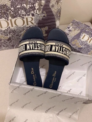 Marinblå Dior sandaler med logga - Snygga marinblå sandaler från Christian Dior med bred textilrem där det står 'Christian Dior Paris' i vitt. Klassisk öppen tå och platt sula. Kommer i originalkartong med Dior-mönstrat papper. Perfekta för en trendig och lyxig look. Finns även i olika färger och storlekar. 