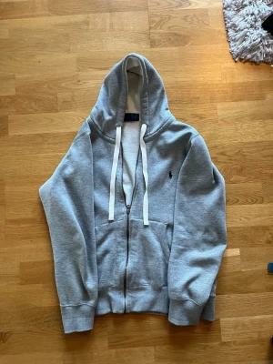 Grå hoodie från Polo Ralph Lauren - Snygg grå zip-hoodie från Polo Ralph Lauren med klassisk broderad logga på bröstet. Hoodien har vita dragsnören, känguruficka och ribbade muddar. Perfekt för en avslappnad och stilren look. Mjuk bomullsblandning för extra komfort. Storlek S.