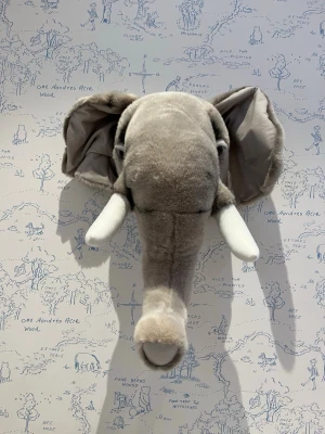 Väggdekoration Elefant från Brigbys - Mjuk väggdekoration i form av ett elefanthuvud i grå plysch med stora öron och vita betar. Perfekt som lekfull inredningsdetalj till barnrum eller som rolig dekoration. Realistisk form och detaljerad design, lätt att hänga upp på väggen.