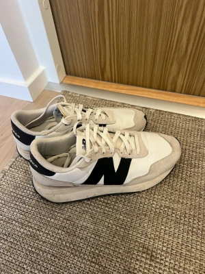 Beige New Balance sneakers med svart N - Snygga sneakers från New Balance i beige mocka och textil med svart N-logga på sidan. Skorna har vita snören, vit ovandel och svart detalj vid hälen. Klassisk låg modell som passar till många olika stilar.