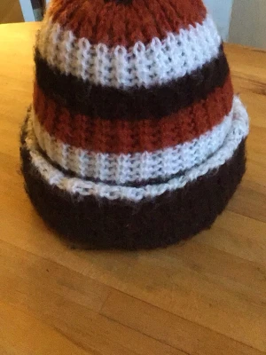 Randig stickad mössa i brunt och orange - Säljer en stickad mössa med breda ränder i brunt, orange och vitt. Mössan är ribbstickad och har en mjuk, lite pösig form. Perfekt för dig som gillar retrovibbar och vill hålla dig varm med stil.