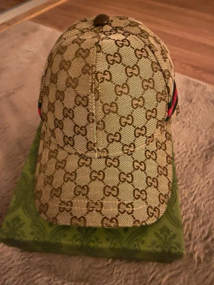 Beige GG keps från Gucci - Säljer en beige keps från Gucci med det klassiska GG-monogrammet över hela kepsen. Kepsen har en böjd skärm och detaljer i rött och grönt på sidan. Tillverkad i canvasmaterial och levereras med originalkartong och dustbag.
