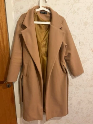 Beige kappa med skärp från Zara - Stilren beige kappa från Zara med klassisk krage, knytband i midjan och två fickor framtill. Kappan har ett mjukt tyg och är helfodrad med ljusgul insida. Perfekt för att lyfta din höst- eller vinterstil.