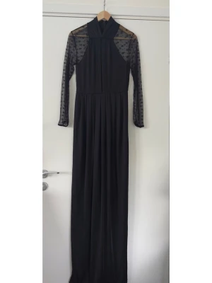 Svart långklänning med meshärmar - helt NY - Elegant svart långklänning med hög krage (med dold dragkedja bak) och långa ärmar i spets. Klänningen har en smickrande passform och passar perfekt för festliga tillfällen. HELT NY!  Hämtas i Stockholm eller skickas mot porto. 