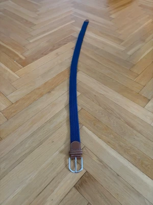 Blått flätat bälte - Snyggt blått bälte med vävd struktur och bruna skinn-detaljer vid spännet. Spännet är i silverfärgad metall och bältet har en klassisk, stilren design som passar till många outfits.