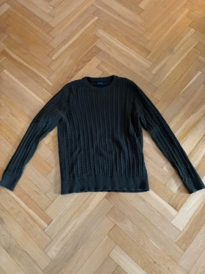 Mörkgrön kabelstickad tröja - Mörkgrön kabelstickad tröja med rund halsringning och långa ärmar. Tröjan har ribbade muddar vid ärmslut och nederkant, vilket ger en snygg och klassisk look. Perfekt för lager-på-lager-stil under kyliga dagar.