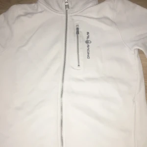 Vit hoodiejacka från Sail Racing - Snygg vit hoodiejacka från Sail Racing med dragkedja framtill och på bröstfickan. Jackan har justerbar huva med snörning och lång ärm. Mjuk insida i bomullsmix och stilren logga på bröstet. Perfekt för chill dagar och streetwear.