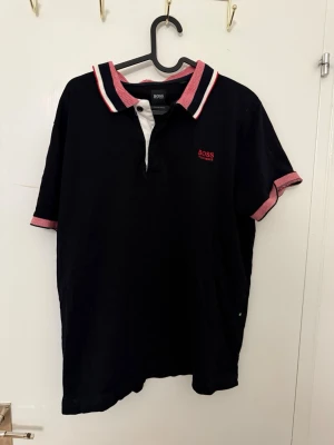 Svart pikétröja från BOSS - Svart pikétröja från BOSS med rosa och vita detaljer på krage och ärmslut. Klassisk polokrage med knappslå och diskret BOSS-logga broderad i rött på bröstet. Tillverkad i mjuk bomull och har korta ärmar.